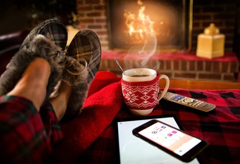 A person's feet wearing slippers with a hot drink next to them on the foot rest and a fire burning in a fireplace in the background